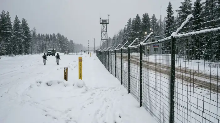 Barrière frontalière sous la neige entre la Finlande et la Russie, gardes-frontières au loin.